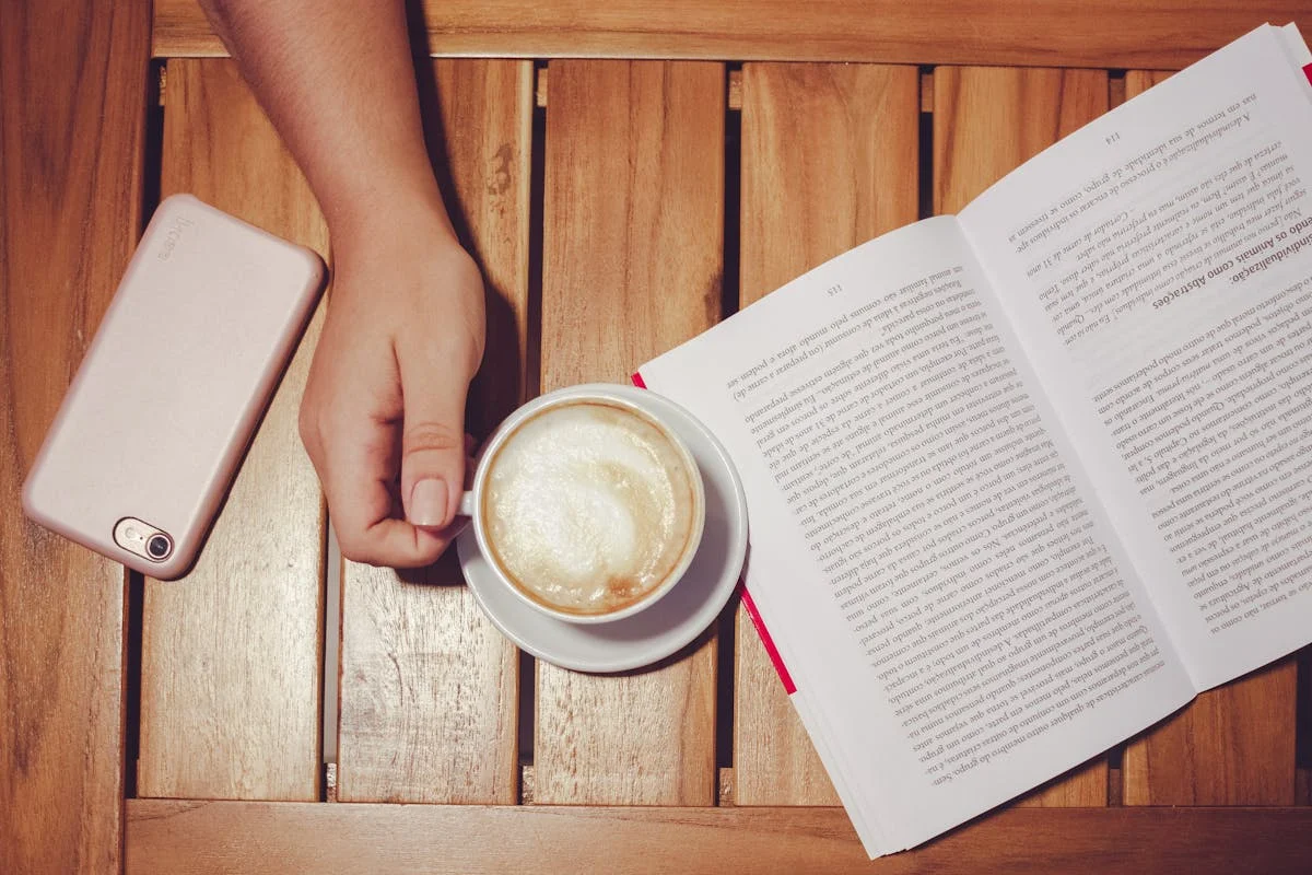Un livre ouvert, une tasse de café et un smartphone posés sur une table en bois.